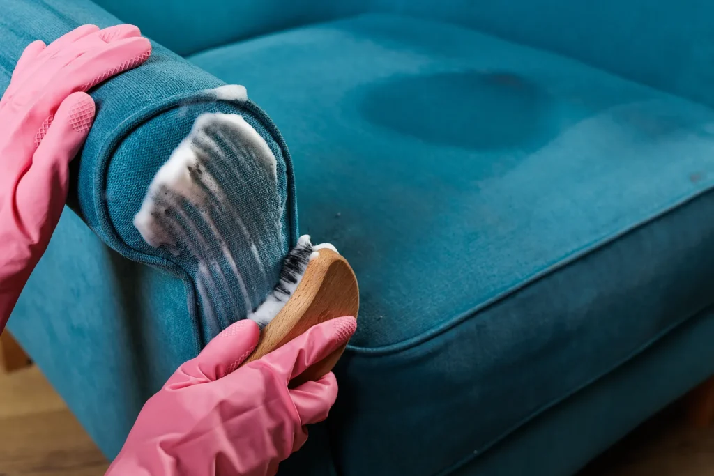 Hands cleaning blue sofa upholstery with a brush and foam